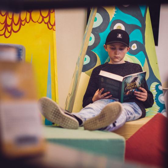 Dreng der sidder og læser i en bog op ad en væg med benene oppe