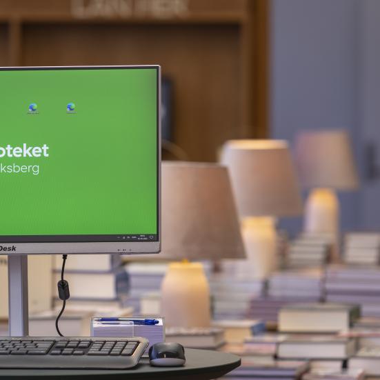 Billede af en computer, med bøger i baggrunden, på Frederiksberg Bibliotek. 