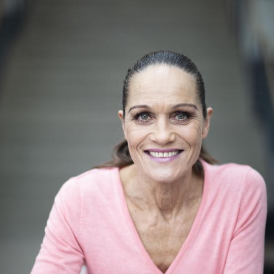 Portrætbillede af Lotte Heise i lyserød trøje stående ved et bord.