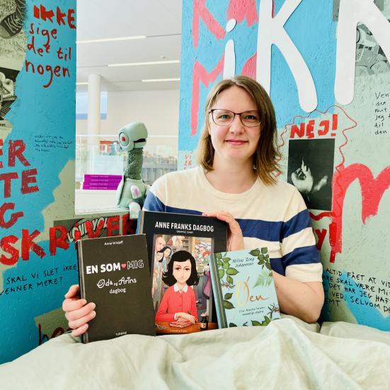 Kvindelig børnekulturformidler siddende i en seng med tre dagbøger udvalgt tiltweens.