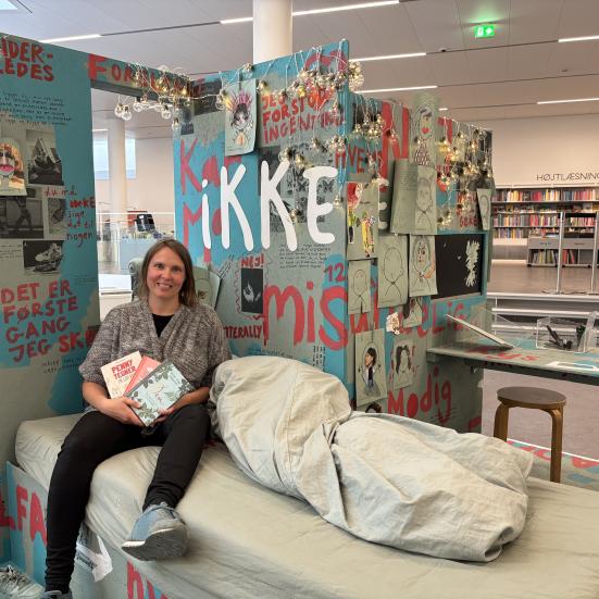 Kulturformidler sidder i seng i installation af teenageværelse med tre dagbøger i hånden.