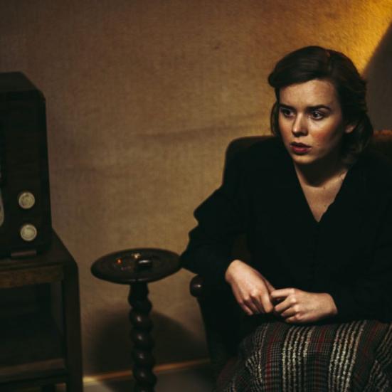 En ung kvinde sidder i en stol foran en gammel radio under besættelsestiden.