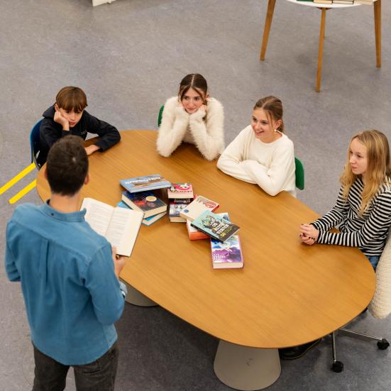 4 unge sidder ved et bord og lytter til en mand der står og læser op af en bog