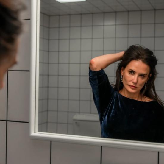 Billede fra filmen The Substance, hvor Demi Moore står foran et spejl og roder sig i håret.