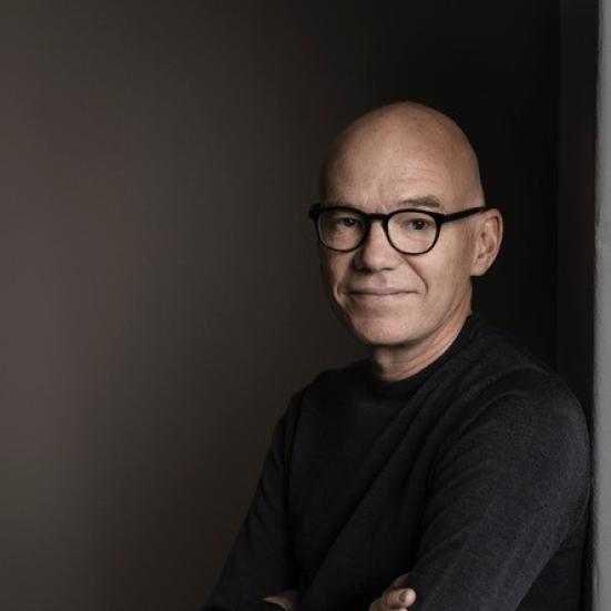 Forfatteren står med sort bluse og mørk baggrund med armene over kors.