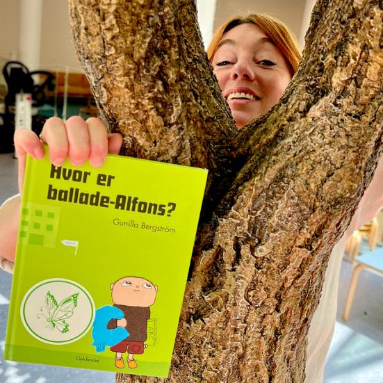 bibliotekar kigger frem fra bag et træ og holder anbefalet bog