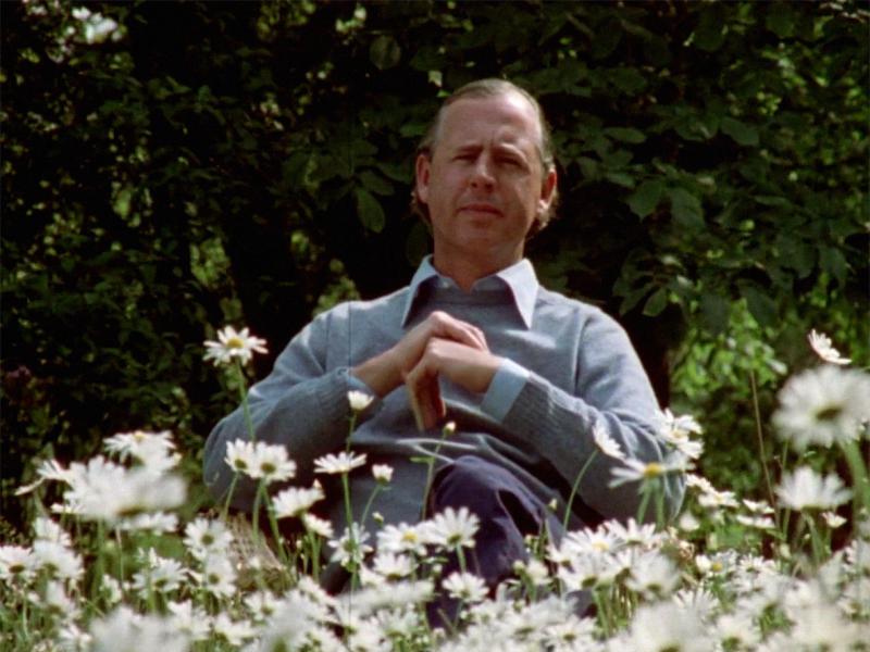 Klaus Rifbjerg siddende i blå skjorte i en have med hvide blomster.