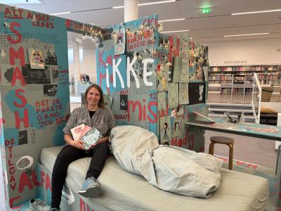 Kulturformidler sidder i seng i installation af teenageværelse med tre dagbøger i hånden.