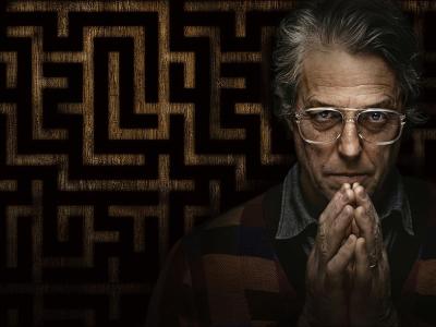 Billede af skuespilleren Hugh Grants ansigt på en mørk baggrund med et mønster. Han folder hænderne foran ansigtet.