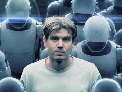 Mand i hvid t-shirt stående blandt en mængde identistiske robotter, der kigger ned