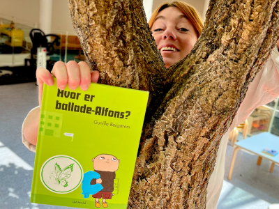 bibliotekar kigger frem fra bag et træ og holder anbefalet bog