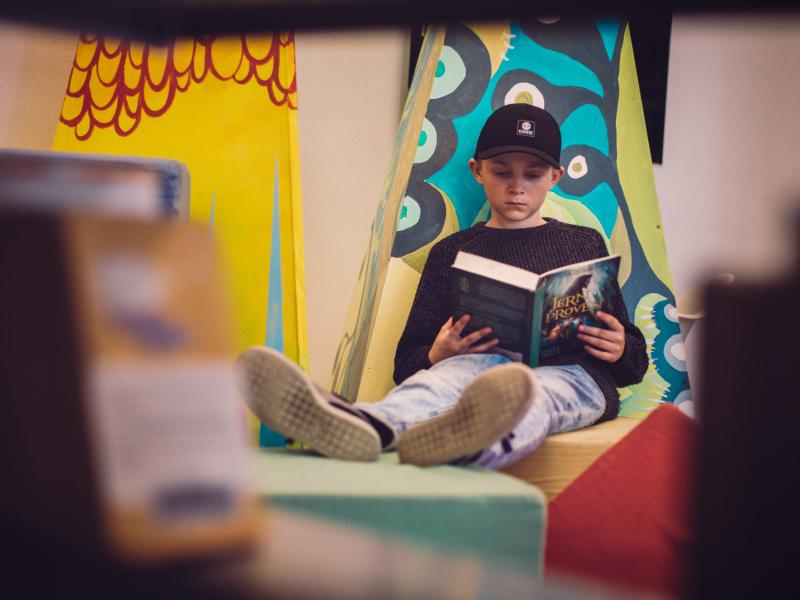 Dreng der sidder og læser i en bog op ad en væg med benene oppe