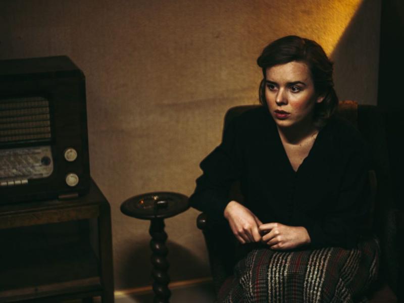 En ung kvinde sidder i en stol foran en gammel radio under besættelsestiden.