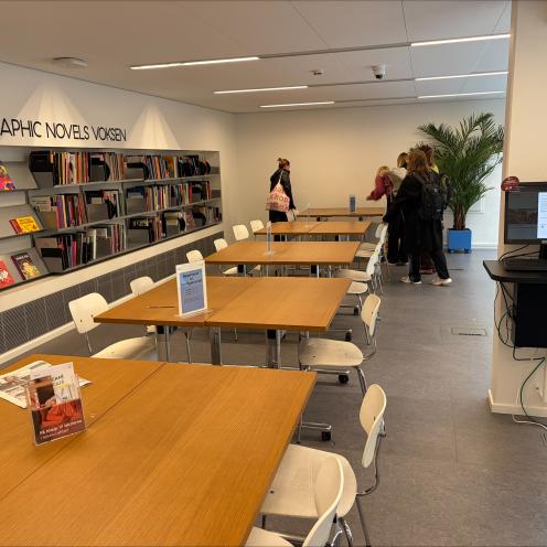 Billede af række med borde og stole og en reol med bøger i baggrunden.
