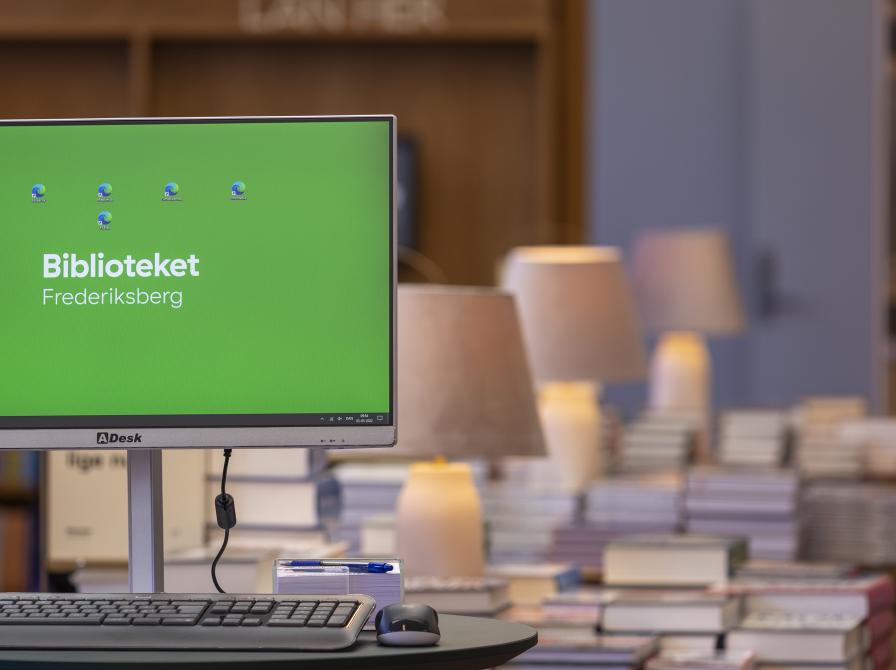 Billede af en computer, med bøger i baggrunden, på Frederiksberg Bibliotek. 