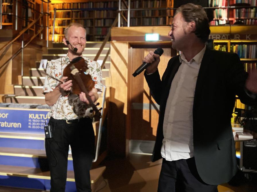 Knud Romer med mikrofon og en bibliotekets mandlige medarbejdere med en violin.