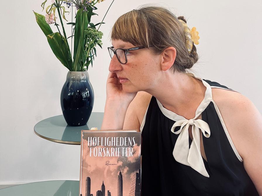 Kvindelig bibliotekar i balkjole siddende ved et bord med bogen stående og en blomstervase i baggrunden.