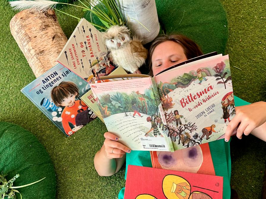 Kulturformidler ligger på græs og læser i billedbog