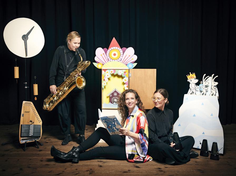 tre musikere, en med en saxofon, en sidder med bogen Skrædderkaninerne og bingebjørnen rejser i tiden.