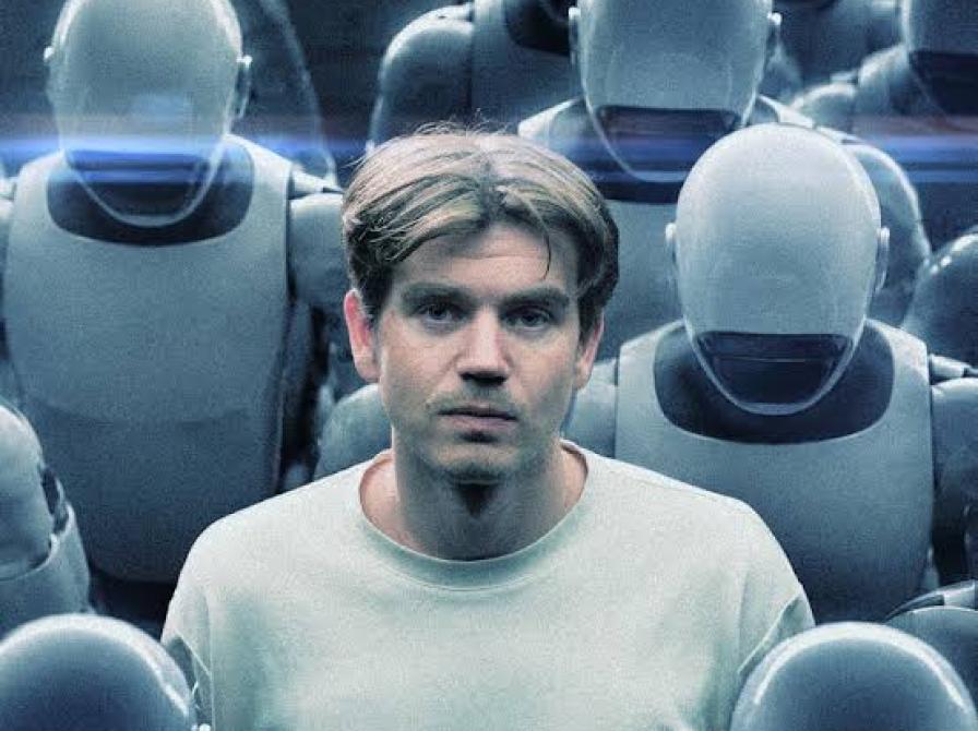 Mand i hvid t-shirt stående blandt en mængde identistiske robotter, der kigger ned