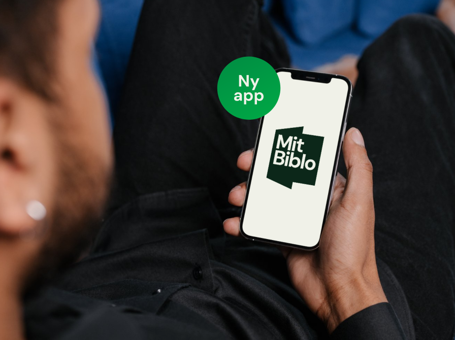 Sløret ansigt  set skråt bagfra, hånd med telefon i fokus, på skærmen "Mit Biblo"-logo. Grøn sticker med teksten "Ny app"
